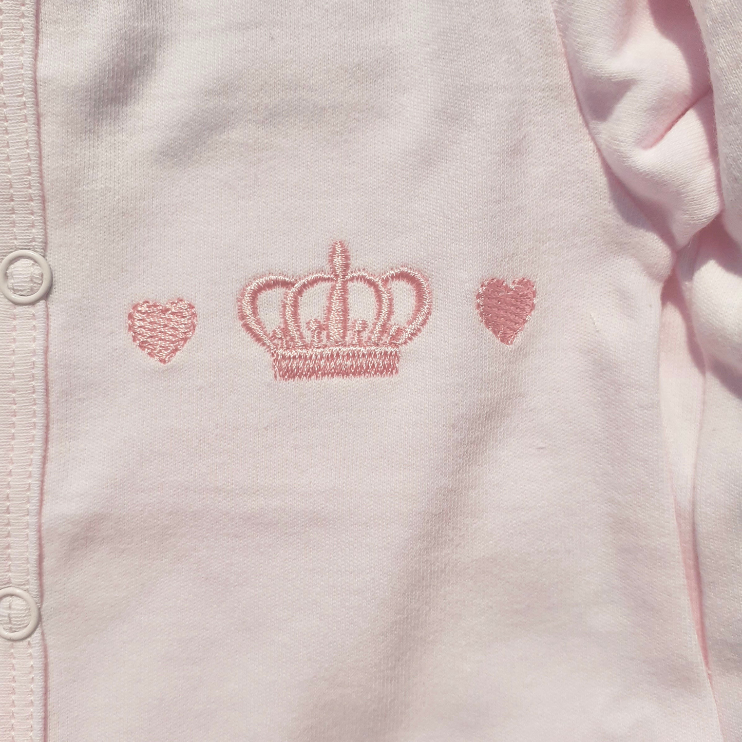 Pink baby outfit with embroidered crown and heart designs on a light pink background
