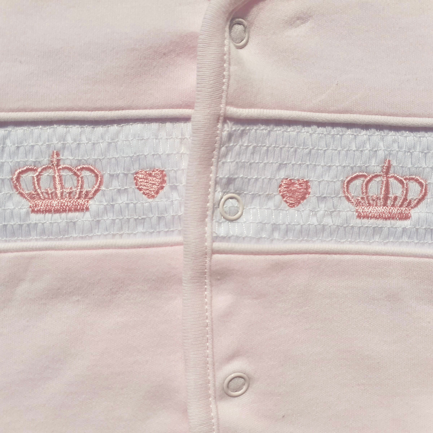 Close-up of a baby outfit with embroidered crowns and hearts on a light pink background.